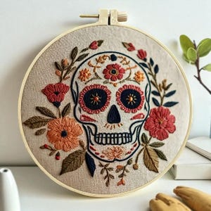 May include: Embroidered skull art in a wooden hoop. The skull is decorated with red, orange, and blue floral designs. The background is a light beige fabric. The embroidery hoop is a light tan color.