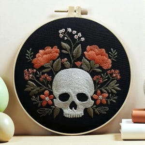 May include: Embroidered skull artwork in a circular frame. The skull is white, encircled by orange and red flowers, green leaves, and small white flowers, set against a black backdrop. The embroidery hoop is beige.