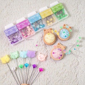May include: A collection of sewing pins and pin cushions. The image shows several clear plastic boxes filled with decorative pins, along with cat-shaped pin cushions and decorative pins with cat and gem-shaped heads. The text "Sewing Pins 80pcs" is visible on the boxes.