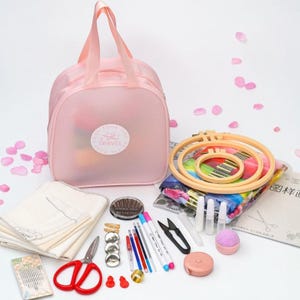May include: A pink travel bag containing embroidery equipment: hoops, threads, needles, scissors, and a tape measure. The bag has a circular logo that reads "Enjoy Your Travel Time". Pink petals are scattered around the items.