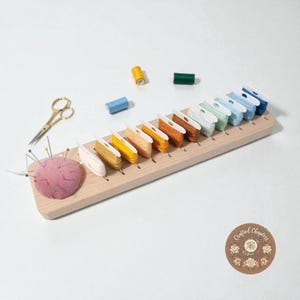 May include: A wooden thread organiser with spools of colourful embroidery floss. A pink pin cushion with needles and gold scissors are also present. Additional spools of thread in yellow, blue, and green are scattered around the organiser.