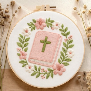 May include: Embroidered hoop art showing a pink book with a green cross, encircled by a wreath of pink and white flowers and green leaves. The embroidery is on white fabric, framed by a wooden hoop.