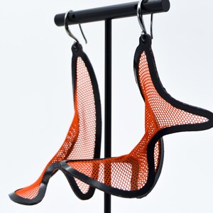 Op de afbeelding: Een paar hangende oorbellen met een feloranje mesh-stoffen binnenkant, omlijst door een effen zwarte rand. De oorbellen hebben een gebogen, organische vorm en hangen aan zilverkleurige metalen haakjes voor een opvallend contrast.