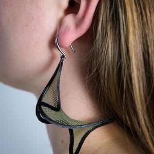 Può includere: Un grande orecchino scultoreo caratterizzato da una cornice scura e solida che delinea un materiale a rete fine di colore verde oliva. L'orecchino ha una forma tridimensionale attorcigliata e pende da un semplice gancio in metallo argentato.