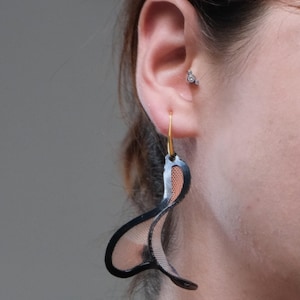 Può includere: Un orecchino pendente caratterizzato da una struttura curva a forma di onda con una cornice nera e un interno in rete fine color rame. L'orecchino è sospeso a un gancio dorato e indossato al lobo, completato da un piccolo orecchino a perno.