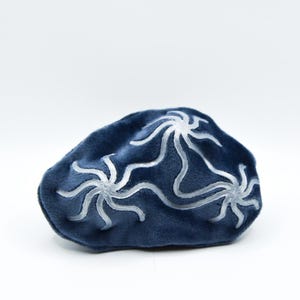 K&ouml;nnte beinhalten: Eine dunkelblaue Schlafmaske mit samtartiger Textur und einem wei&szlig;en, stilisierten Blumenmuster. Die Maske ist oval und vor einem wei&szlig;en Hintergrund platziert. Das Design ist symmetrisch und scheint handgezeichnet zu sein.