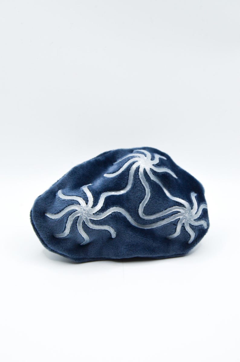 K&ouml;nnte beinhalten: Eine dunkelblaue Schlafmaske mit samtartiger Textur und einem wei&szlig;en, stilisierten Blumenmuster. Die Maske ist oval und vor einem wei&szlig;en Hintergrund platziert. Das Design ist symmetrisch und scheint handgezeichnet zu sein.