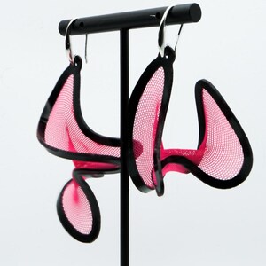 Può includere: Un paio di orecchini pendenti caratterizzati da una rete rosa acceso racchiusa in una cornice nera solida. Gli orecchini presentano una forma curva e astratta e sono sospesi a ganci in metallo color argento dal design leggero.