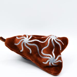 K&ouml;nnte beinhalten: Ein braunes, pl&uuml;schiges, dreieckiges Kissen mit samtiger Textur. Das Kissen hat ein silbernes, stilisiertes Blumenmuster. Das Kissen befindet sich vor einem wei&szlig;en Hintergrund.
