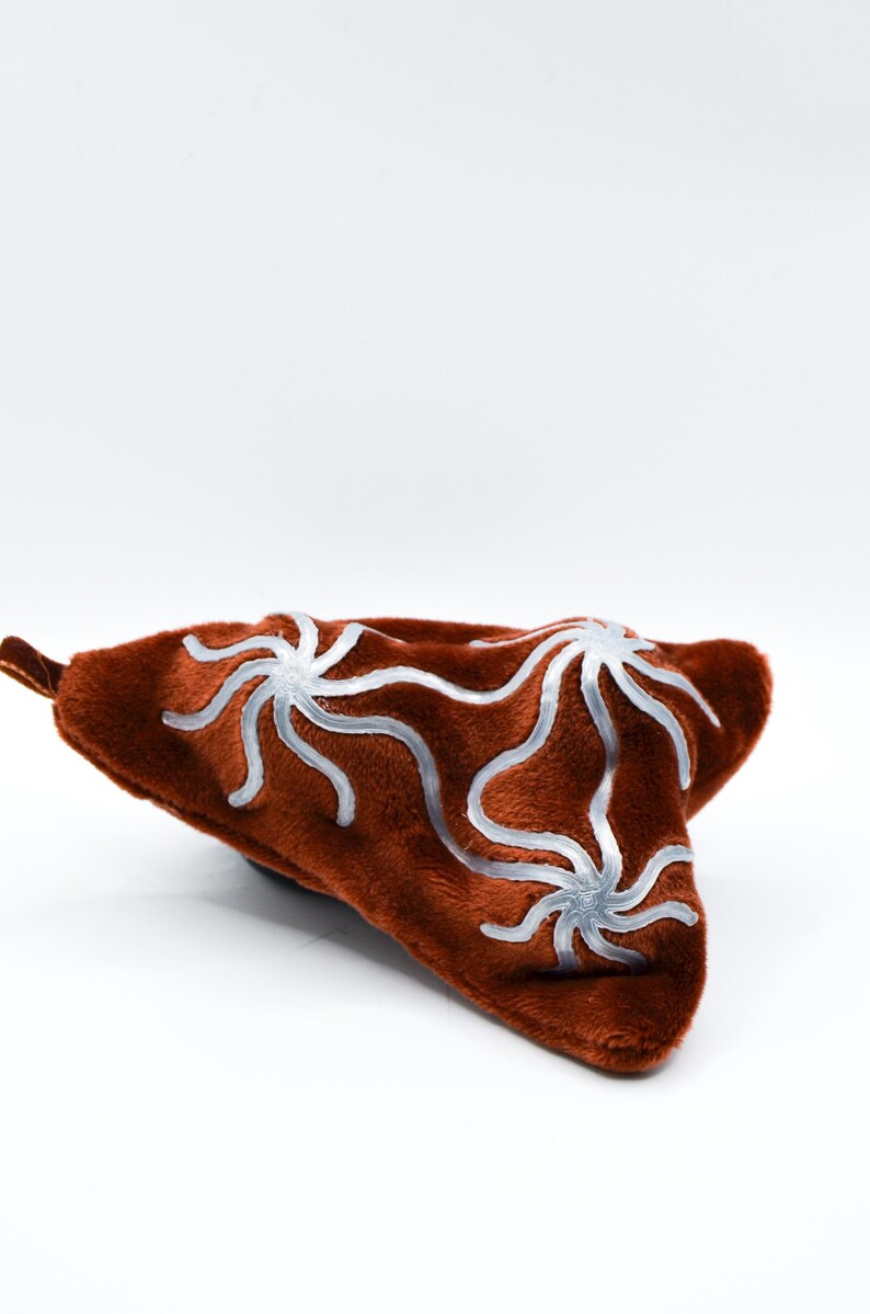 K&ouml;nnte beinhalten: Ein braunes, pl&uuml;schiges, dreieckiges Kissen mit samtiger Textur. Das Kissen hat ein silbernes, stilisiertes Blumenmuster. Das Kissen befindet sich vor einem wei&szlig;en Hintergrund.