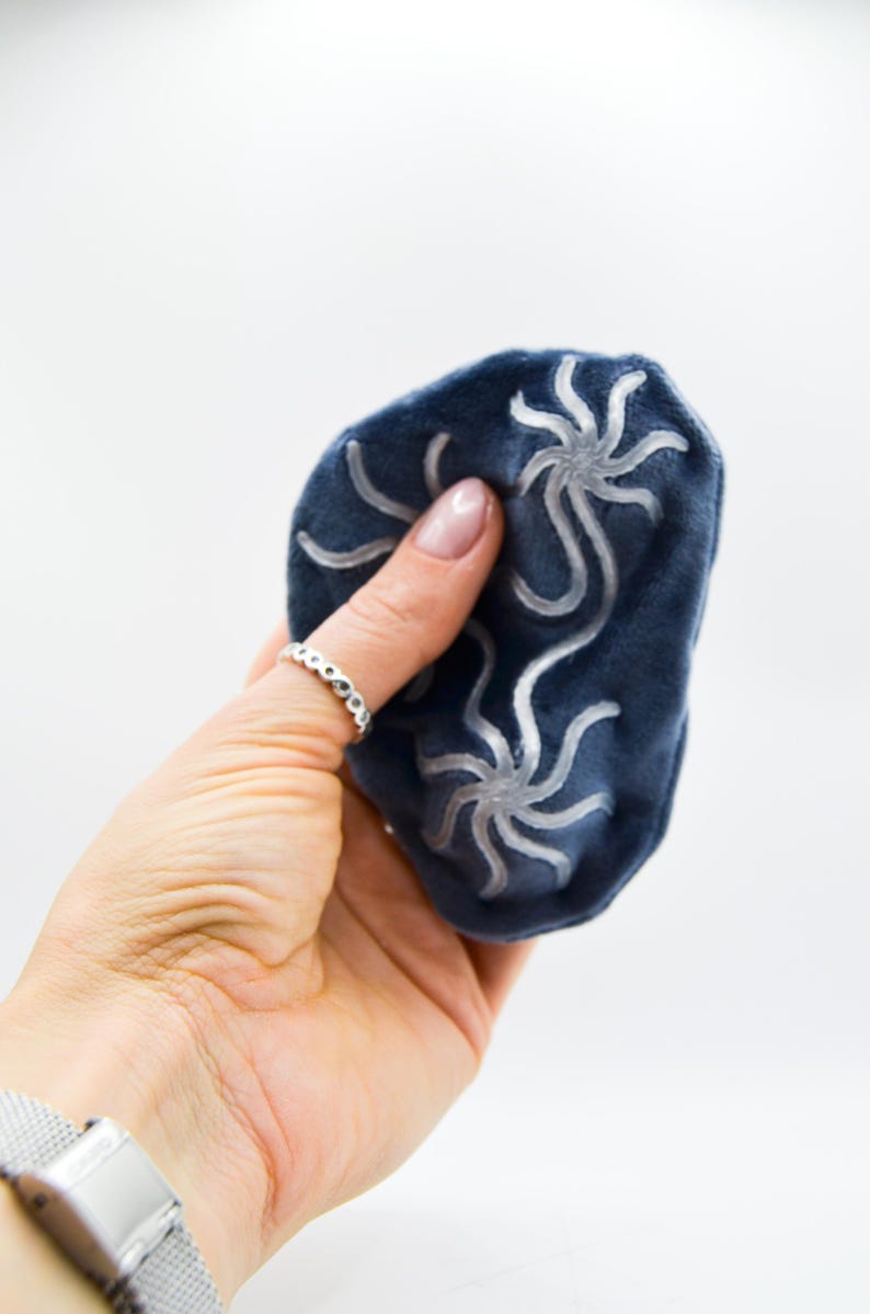 K&ouml;nnte beinhalten: Eine marineblaue Augenmaske aus Samt mit einem stilisierten silbernen Blumendesign. Die Maske wird in einer Hand gehalten, und ein silberner Ring ist an einem Finger zu sehen. Der Hintergrund ist wei&szlig;.