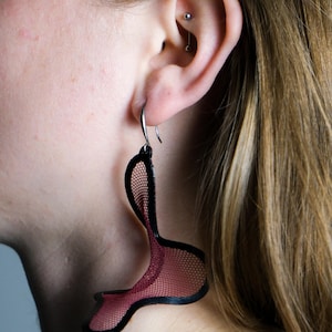 May include: A close-up of a dark red and black earring. The earring has a net-like design with a black outline. The earring is hanging from a silver hook. The earring is worn on the ear.