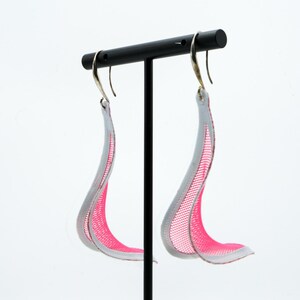 Op de afbeelding: Een paar hangende oorbellen met een gebogen, golfachtige structuur gemaakt van fel neonroze gaasstof met een witte rand. De oorbellen hangen aan zilverkleurige metalen haken, wat zorgt voor een vloeiend en langwerpig silhouet.
