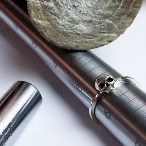 Könnte beinhalten: Ein silberner Totenkopfring auf einem Metall-Ringmaß. Das Ringmaß ist mit Zahlen von 3 bis 9 gekennzeichnet.