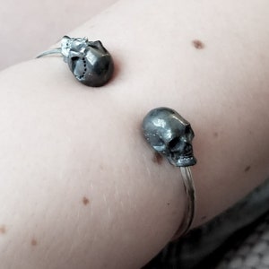May include: A silver bracelet with two skull charms. The bracelet is on a person's arm.