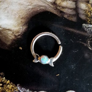 May include: Silver septum ring with a crescent moon design and an iridescent opal gemstone.