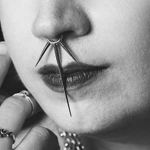 May include: A close-up of a person's face with a nose ring featuring a starburst design. The person is wearing multiple rings on their fingers and a silver chain necklace.
