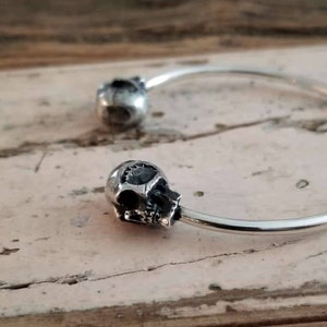 May include: A silver bangle bracelet with two skull-shaped charms. The bracelet is open and has a simple design.