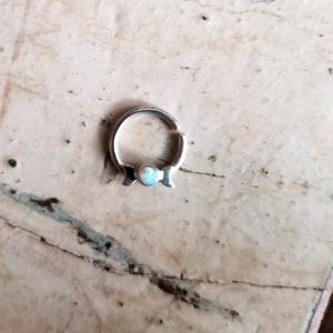 May include: A silver hoop earring with a small opal gemstone in the center. The hoop is open and has a hinged closure.