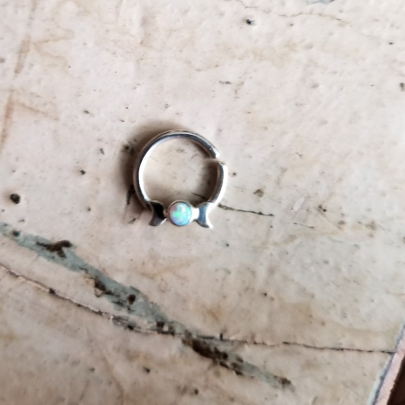 May include: A silver hoop earring with a small opal gemstone in the center. The hoop is open and has a hinged closure.