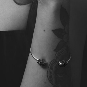 May include: A silver skull cuff bracelet with two skull accents. The bracelet is worn on a person's wrist with a floral tattoo.
