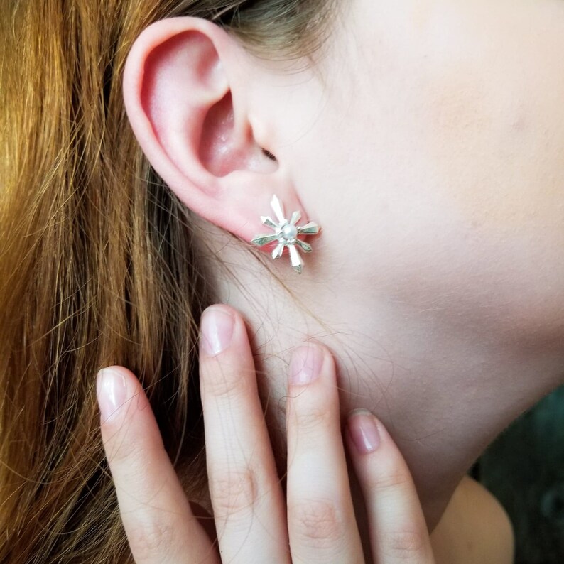 May include: Silver starburst earrings with a pearl centre. The earrings are worn by a person with red hair.