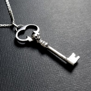 Op de afbeelding: Een zilveren sleutel hanger ketting met een schedel ontwerp. De sleutel heeft een grote, sierlijke boog en een lange, rechte schacht.