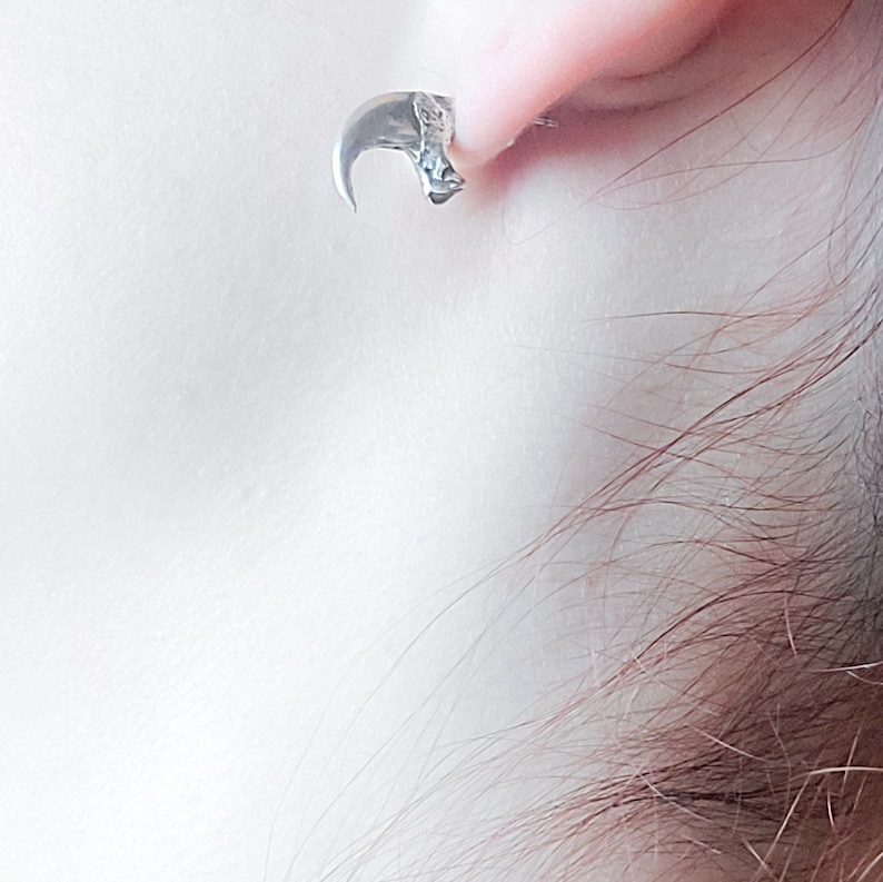 May include: Silver claw earring with a detailed design. The earring is worn in the earlobe of a person.