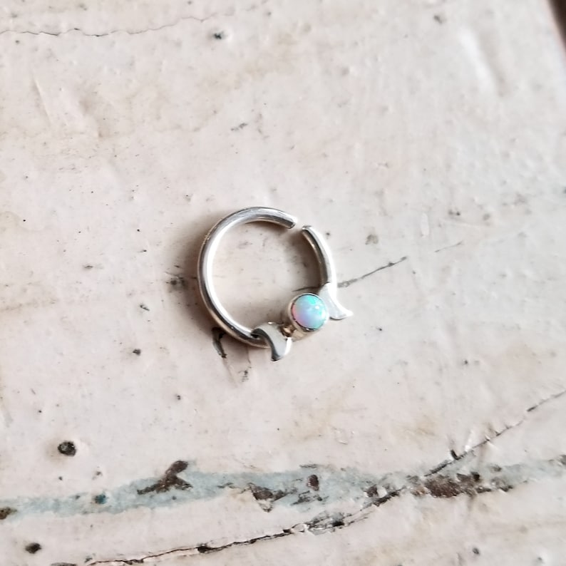 May include: Silver hoop earring with a crescent moon design and a small opal gemstone.