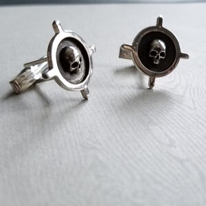 May include: Two silver cufflinks with a skull design in the center. The skull is raised and has a detailed design. The cufflinks are in the shape of a compass with four arms.