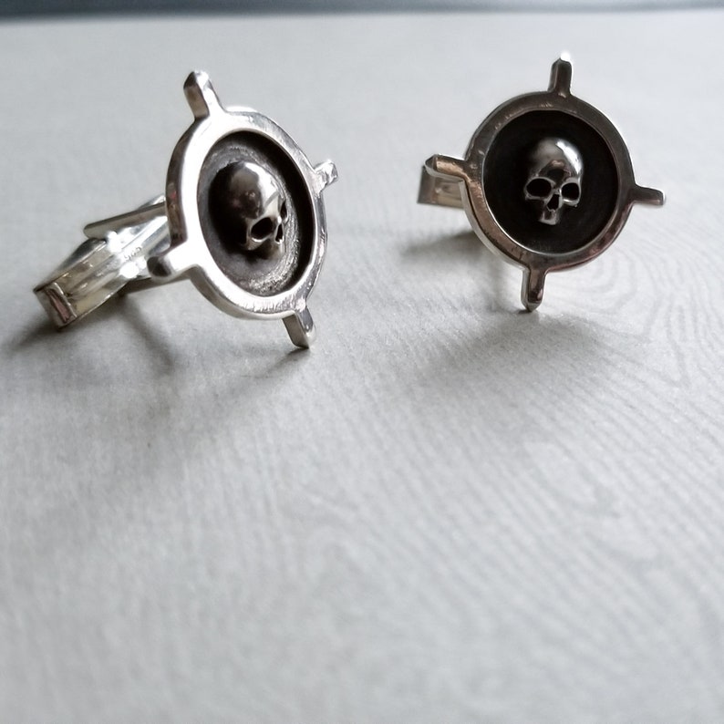 May include: Two silver cufflinks with a skull design in the center. The skull is raised and has a detailed design. The cufflinks are in the shape of a compass with four arms.