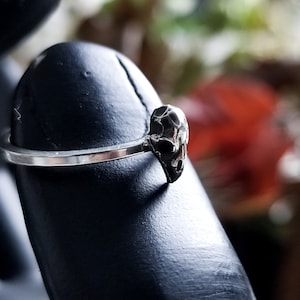 May include: A silver ring with a skull-shaped design. The ring is on a black ring display stand.