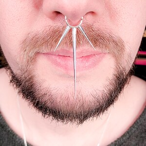 May include: A close-up of a person's face with a silver septum ring with five spikes. The ring is pierced through the septum and the spikes extend down towards the person's lip.
