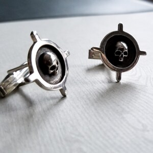 May include: A pair of silver cufflinks with a skull design in the center of each cufflink. The skull is surrounded by a circle with four arms extending out from the circle.