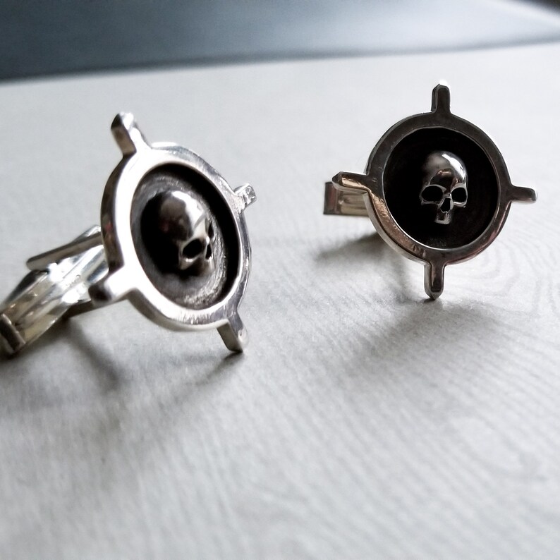May include: A pair of silver cufflinks with a skull design in the center of each cufflink. The skull is surrounded by a circle with four arms extending out from the circle.