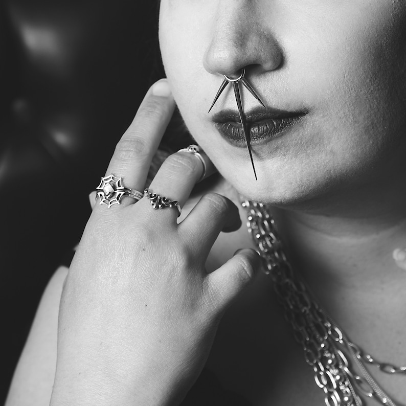 May include: A close-up of a person's face with a nose ring featuring a starburst design. The person is wearing multiple rings on their fingers and a silver chain necklace.