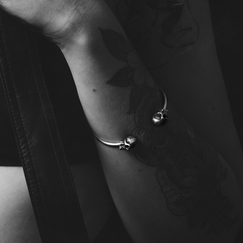 May include: A silver bracelet with two skull charms on a person's arm. The bracelet is on the lower part of the arm, just above the wrist.