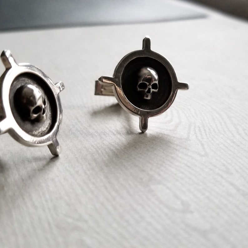 May include: A pair of silver cufflinks with a skull design in the center of each. The skull is surrounded by a circle with four points extending out from the circle.