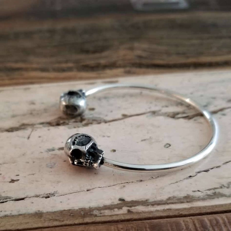 May include: A silver bangle bracelet with two skull-shaped charms. The bracelet is open and has a simple design.