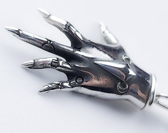 May include: A silver hand charm with detailed finger and knuckle designs. The hand is in a gesture with the index and middle fingers extended and the other fingers curled.