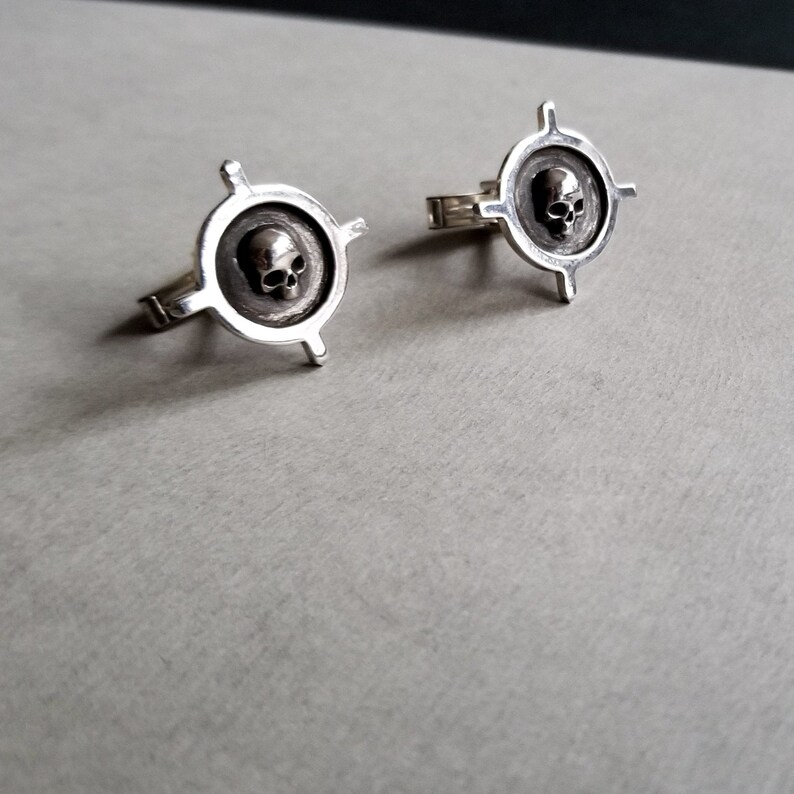 May include: Two silver rings with a skull design in the center of each ring. The rings are in the shape of a target with four arms.