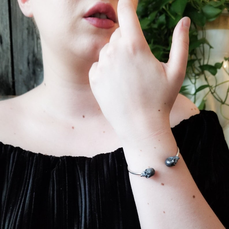 May include: A silver cuff bracelet with two black skull charms. The bracelet is on a person's wrist.