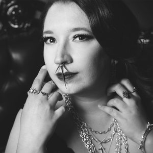 May include: A woman with dark hair and dark eyes is wearing a variety of silver necklaces and rings. She has a nose ring with multiple spikes.