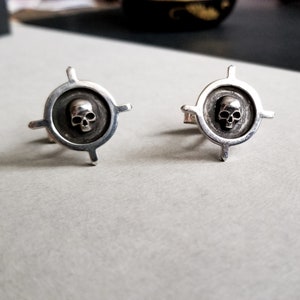 May include: A pair of silver cufflinks with a skull design in the center of each. The skull is surrounded by a circular design with four points extending out from the circle.