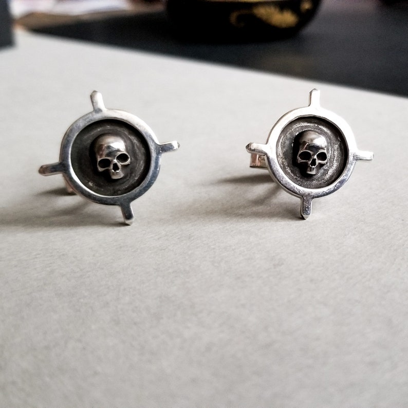 May include: A pair of silver cufflinks with a skull design in the center of each. The skull is surrounded by a circular design with four points extending out from the circle.