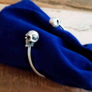May include: A silver skull cuff bracelet with two skull accents. The bracelet is on a blue velvet surface.