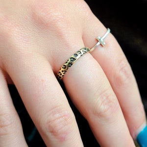 May include: A gold ring with a leopard print design on a person's finger. The ring is worn on the ring finger of the left hand. A silver ring with a cross design is worn on the middle finger of the same hand.