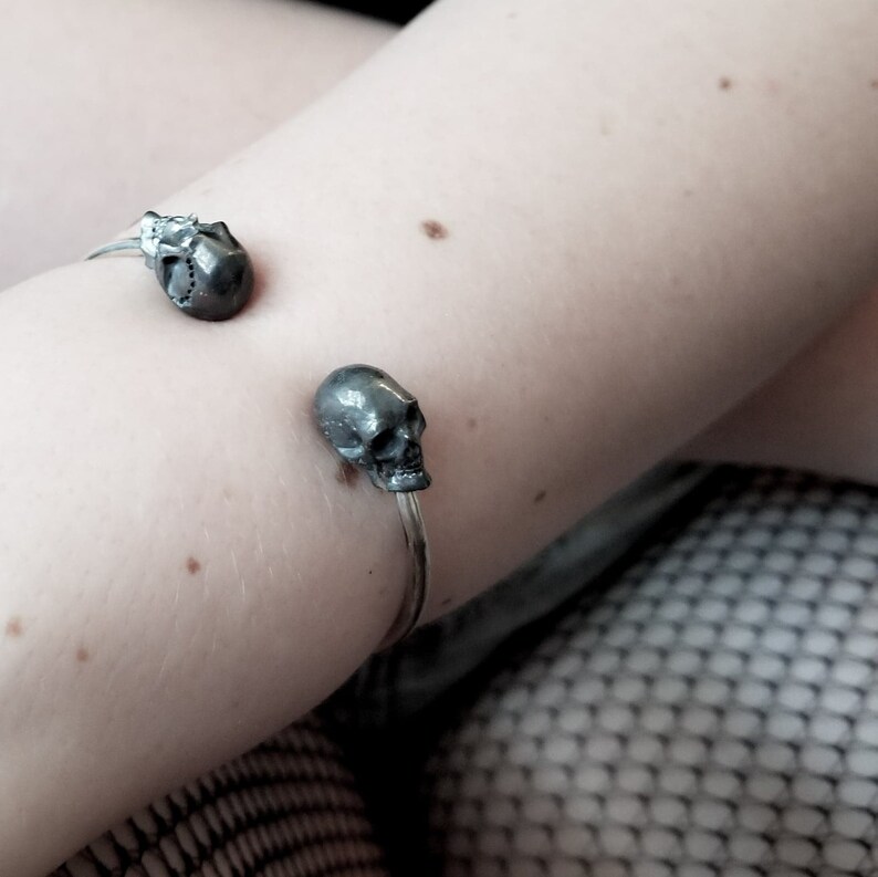 May include: A silver bracelet with two skull charms. The bracelet is on a person's arm.