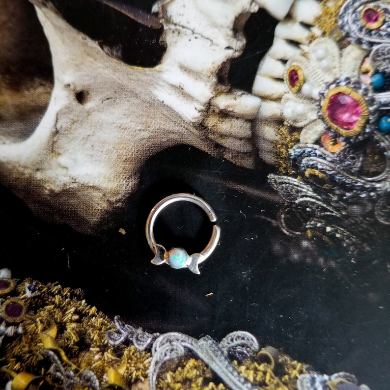 May include: Silver septum ring with a crescent moon design and an iridescent opal gemstone.