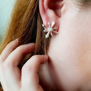 May include: Silver snowflake earrings with a pearl in the centre. The earrings are shown on a person's ear.